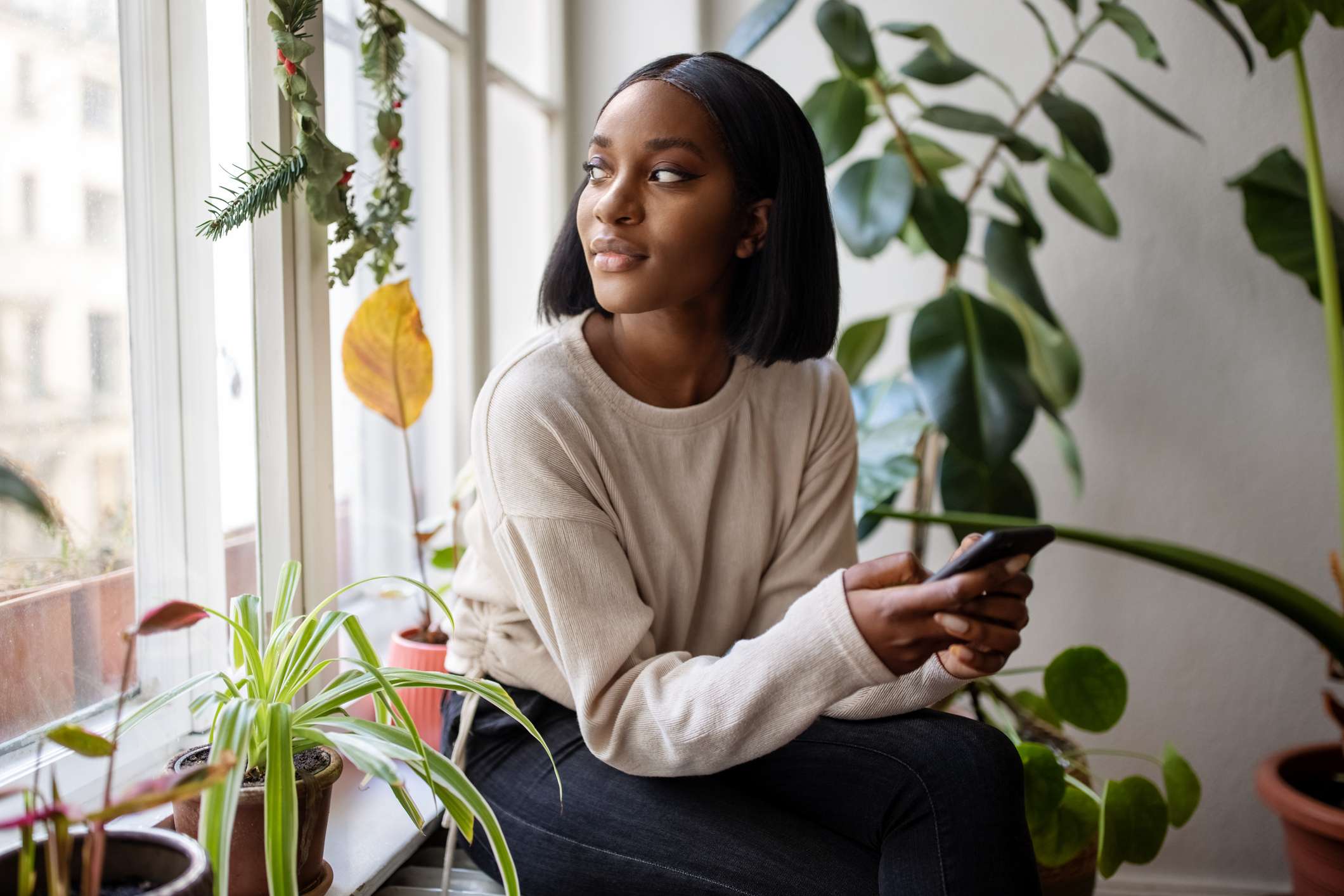 Woman-sitting-on-the-windowsill-with-mobile-phone-and-looking-away-how-to-sleep-earlier